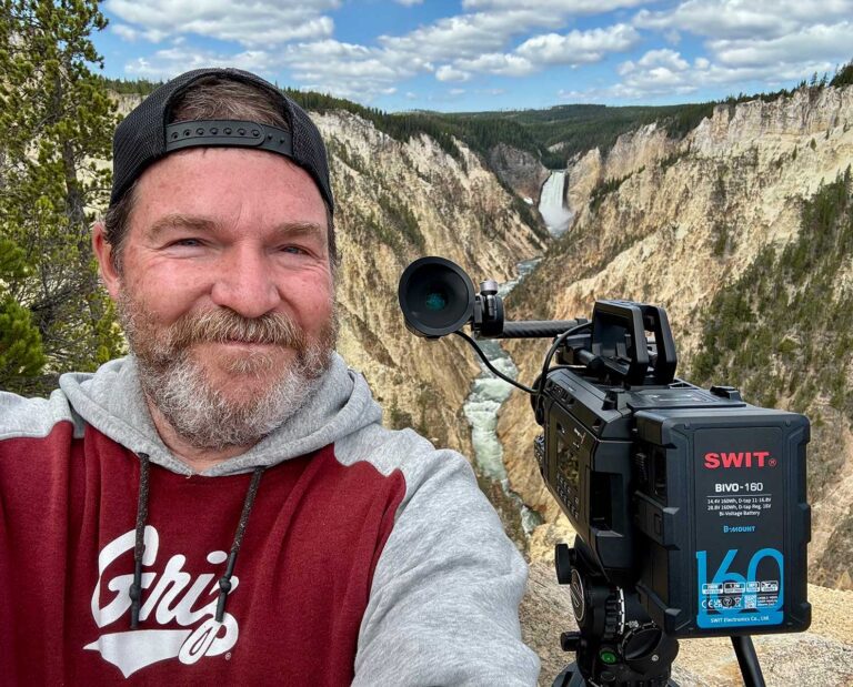 mike-dreesman-with-camera Mike Dreesman, Photographer and Videographer standing next to a video camera with a beautiful mountain vista in the background